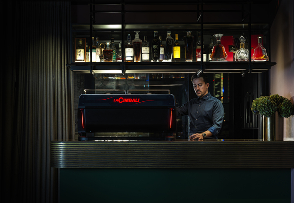 Barista operating a La Cimbali M200 espresso machine in a modern coffee bar with a stylish backlit shelf of liquor bottles